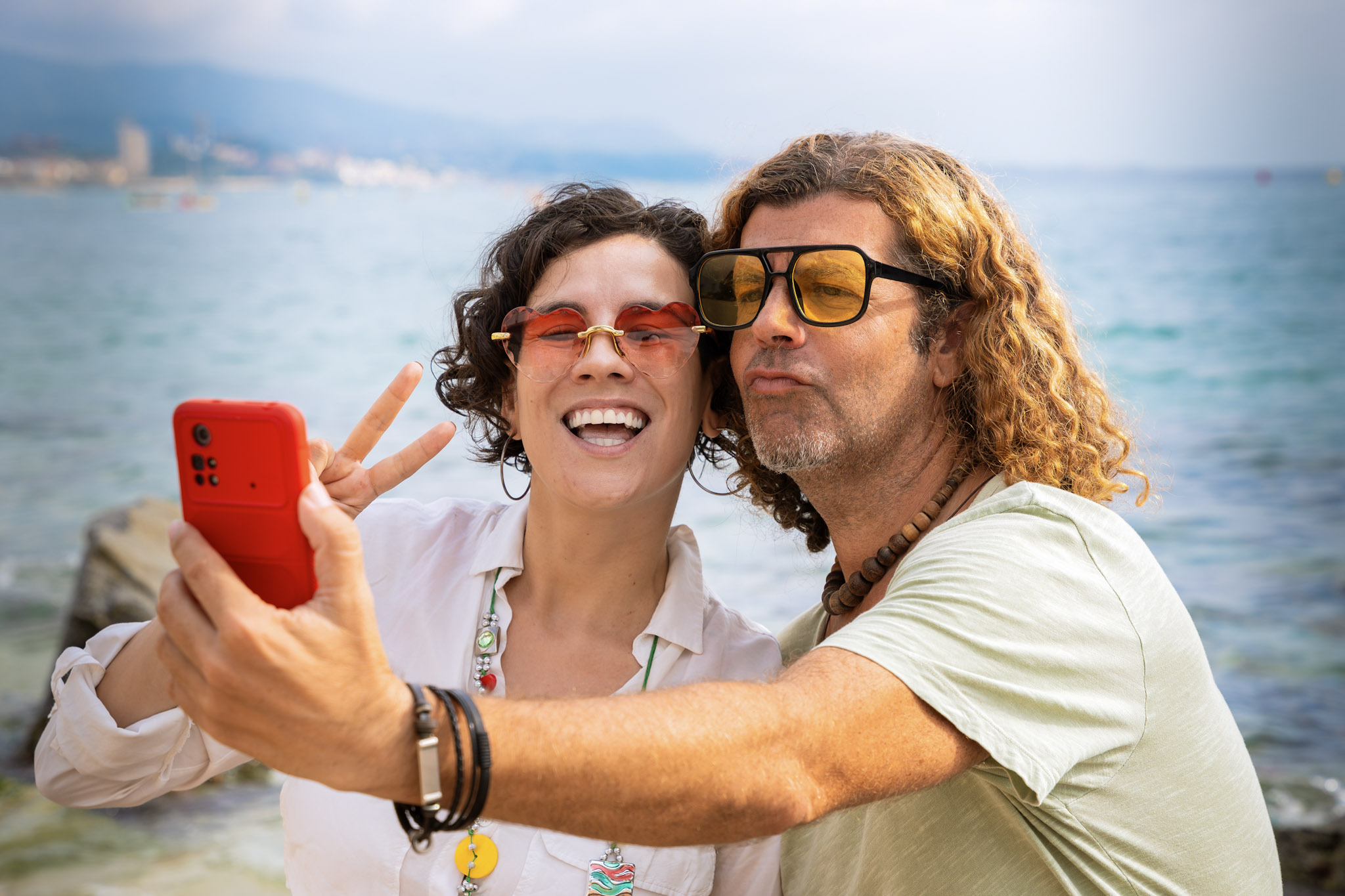 stock image of a woman and man, both wearing sunglasses, taking a selfie using a smartphone while posing by the waterside