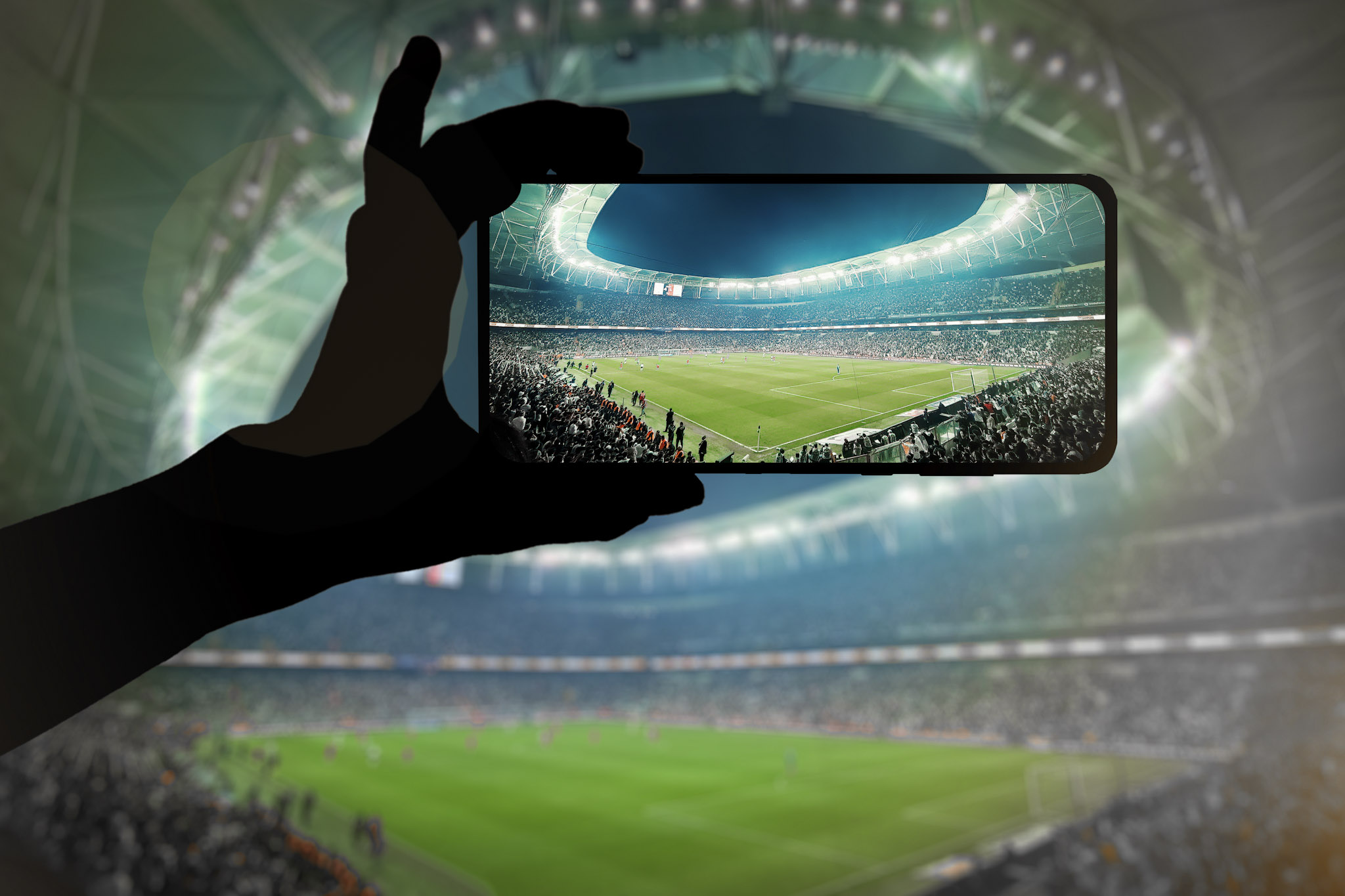 a stock photo of a sports fan's hand holding a mobile phone up to photograph a football game in a stadium [Adobe Stock]